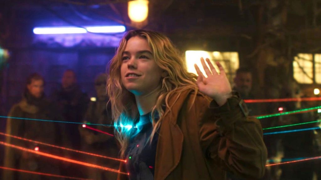 A smiling blonde young woman raises her hand while standing amid colorful laser beams in a lively, neon-lit bar with patrons watching in the background.