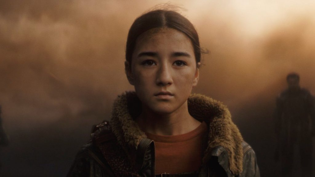 A young girl with a dirt-smudged face and dark hair tied back stands solemnly amid a smoke-filled battlefield, wearing a fur-lined jacket as blurred figures linger in the background.