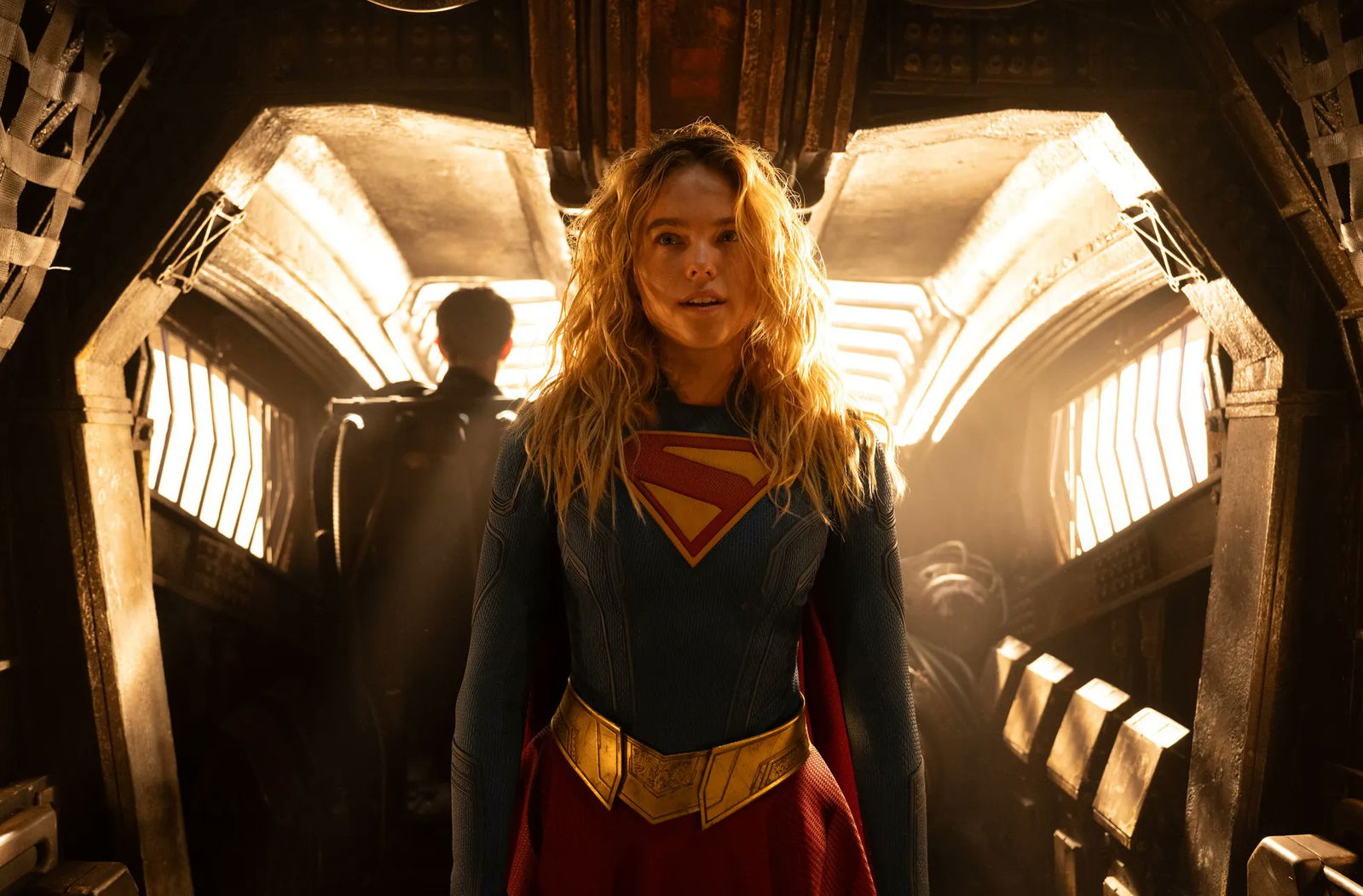 Milly Alcock as Supergirl stands inside a warmly lit Kryptonian spacecraft, wearing her blue-and-red suit as sunlight streams through the ship's interior.