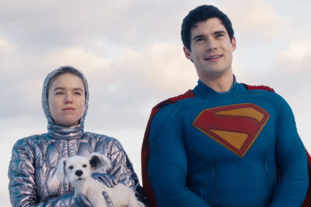 Supergirl stands beside Superman while holding Krypto the Superdog, as the two Kryptonian heroes look toward the horizon