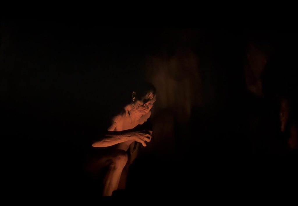 The cyclops creature crouches in near-total darkness, its face and outstretched hand illuminated by the glow of a nearby fire in a scene from The Odyssey.