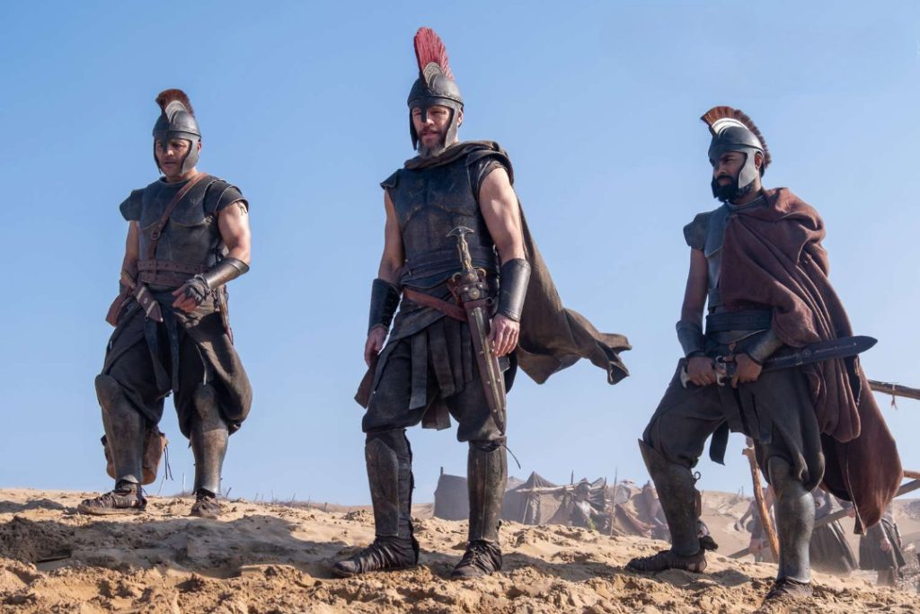 Three armored Greek warriors wearing bronze helmets stand on a sandy shoreline with weapons at their sides in a scene from Christopher Nolan's The Odyssey.