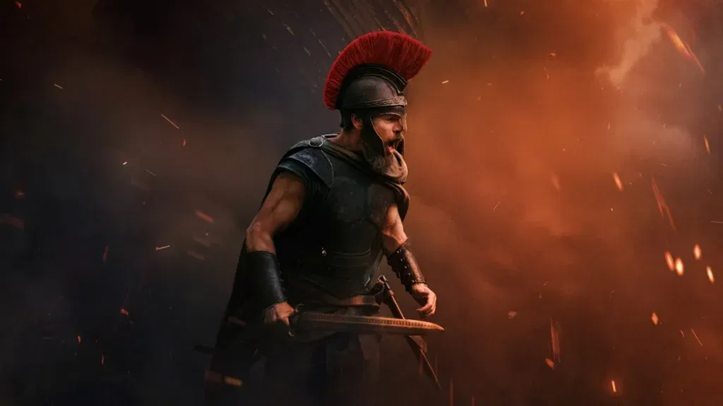 A Greek warrior in black armor and a red-crested helmet brandishes a sword and shouts amid fire, smoke, and flying embers in a battle scene from Christopher Nolan's The Odyssey.
