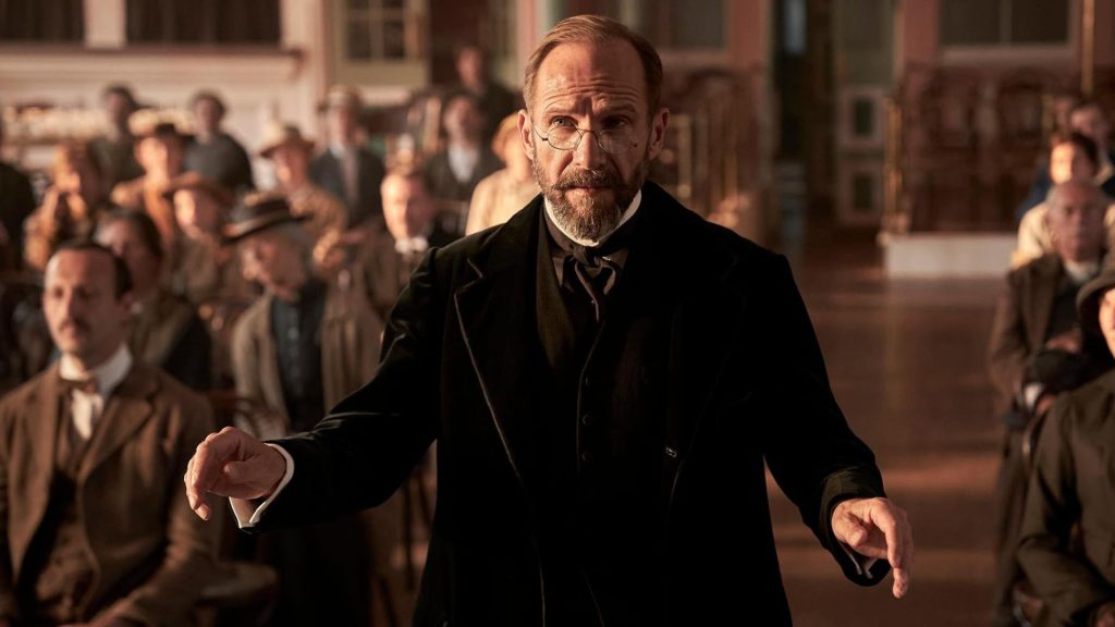 Ralph Fiennes conducts a choir inside a church in a scene from The Choral (2025).