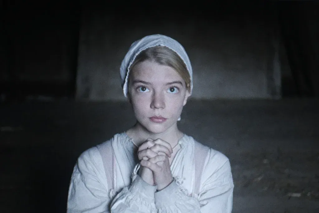 Anya Taylor-Joy as Thomasin clasps her hands in prayer while staring ahead inside a dimly lit farmhouse in The Witch (2015).