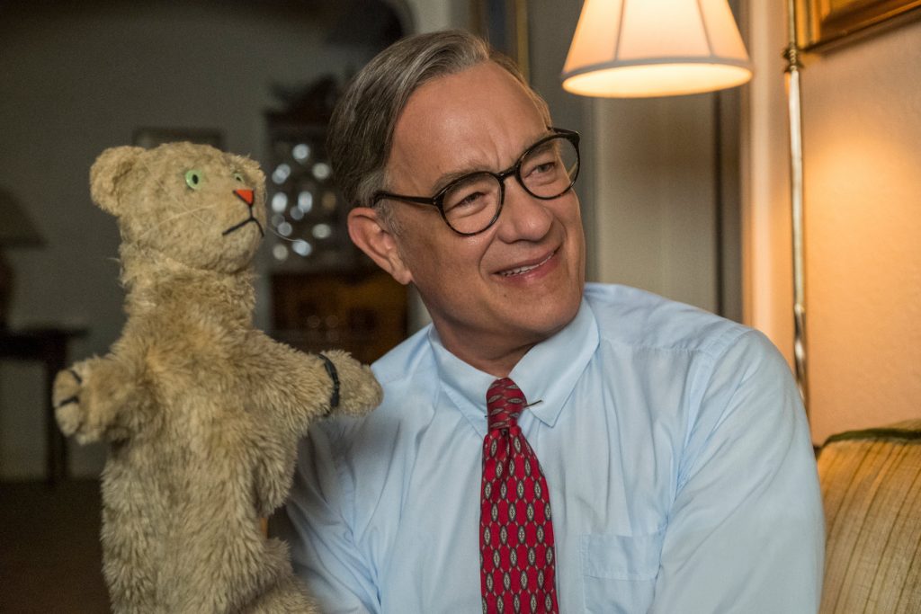 Tom Hanks as Fred Rogers smiles while holding the iconic Daniel Striped Tiger puppet inside a warmly lit living room in A Beautiful Day in the Neighborhood (2019).