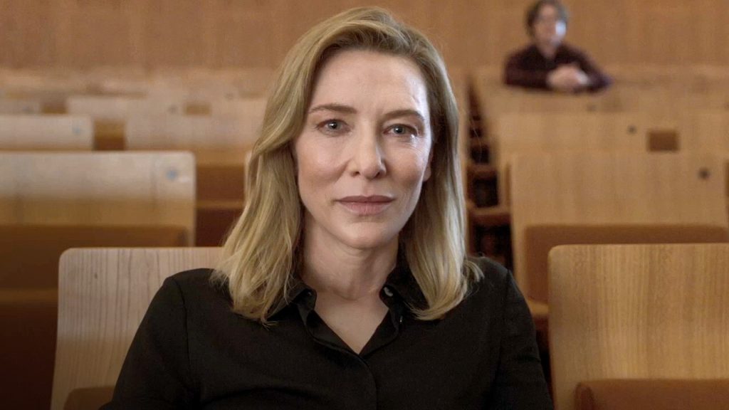 Cate Blanchett as renowned conductor Lydia Tár sits in an empty concert hall, staring ahead with a composed but introspective expression in TÁR (2022).