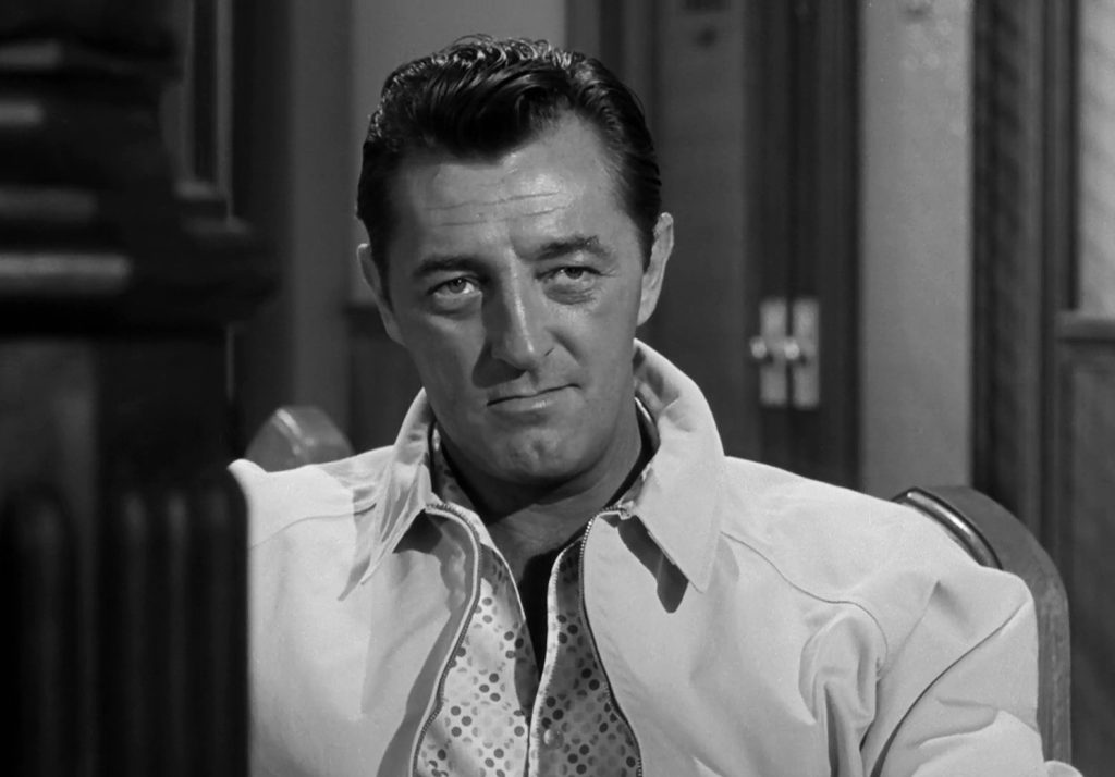 Robert Mitchum as Max Cady in a black-and-white still from Cape Fear, staring coldly at the camera in an unzipped jacket.