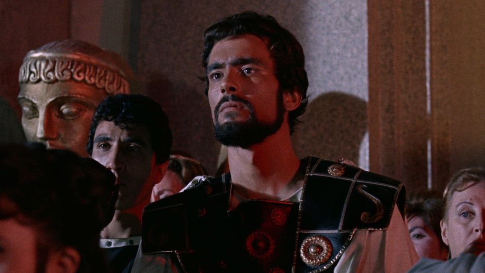 Jason stands in ceremonial Greek armor with a determined expression as onlookers gather around him inside a grand hall in Jason and the Argonauts (1963). 