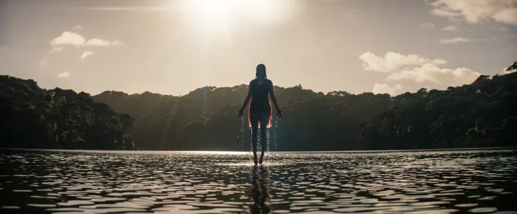 A silhouetted woman emerges from the middle of a calm lake with water dripping from her hands as golden sunlight pours through the clouds behind her, creating an eerie yet serene contrast in Evil Dead Burn.