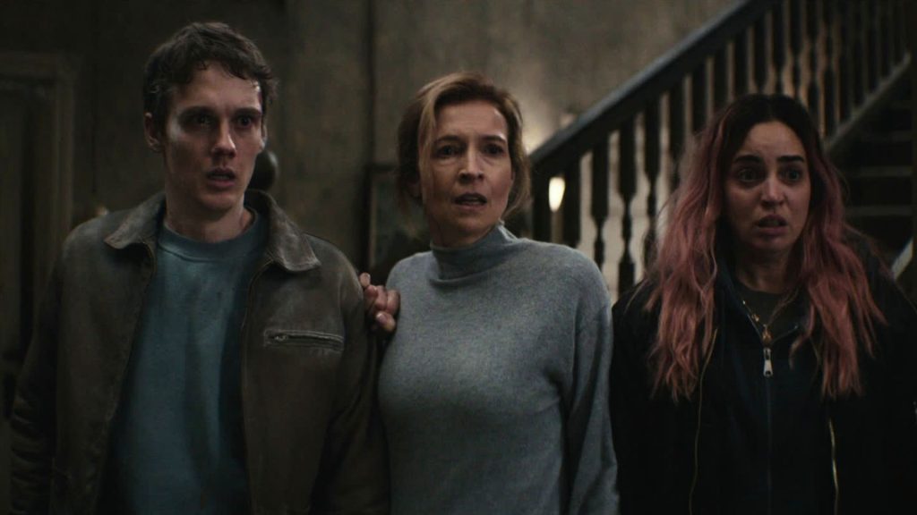 Three terrified family members stand shoulder to shoulder inside a dimly lit house, staring in horror at an unseen threat as a wooden staircase looms behind them in Evil Dead Burn.