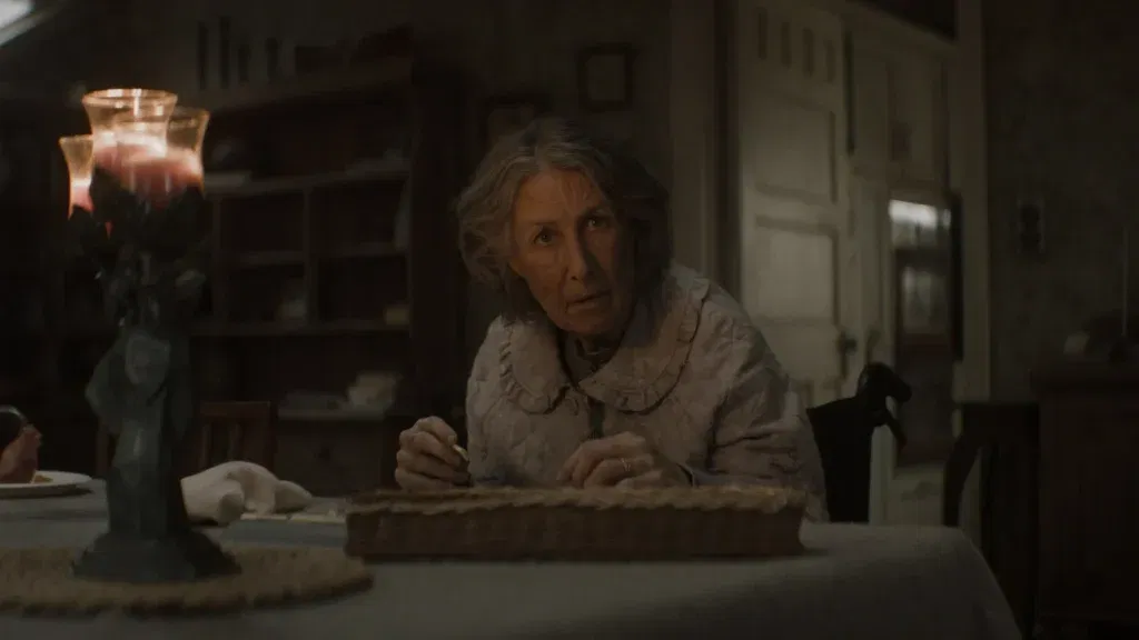 An elderly woman sits at a dining table inside a dimly lit farmhouse, looking up with a worried expression as candlelight flickers beside her in Evil Dead Burn.