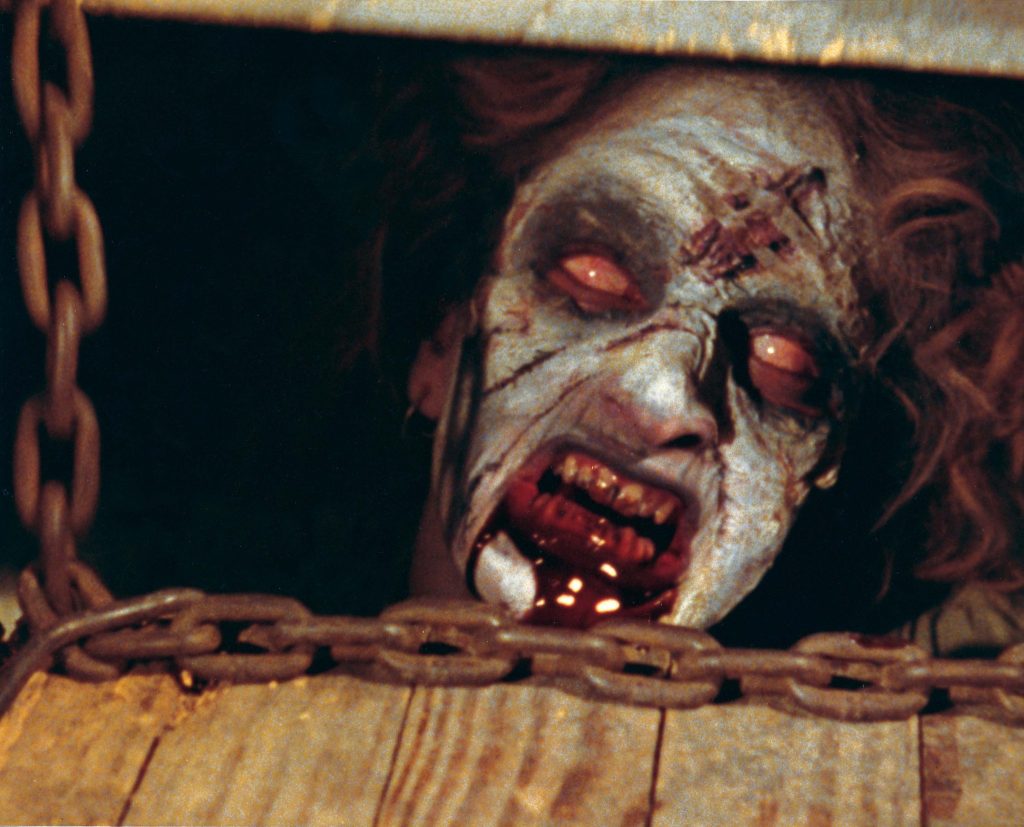 A close-up of a grotesque, pale-faced possessed woman with glowing white eyes, bloodied teeth, and decaying skin glaring from behind heavy chains and a wooden trapdoor in a dark, claustrophobic setting.