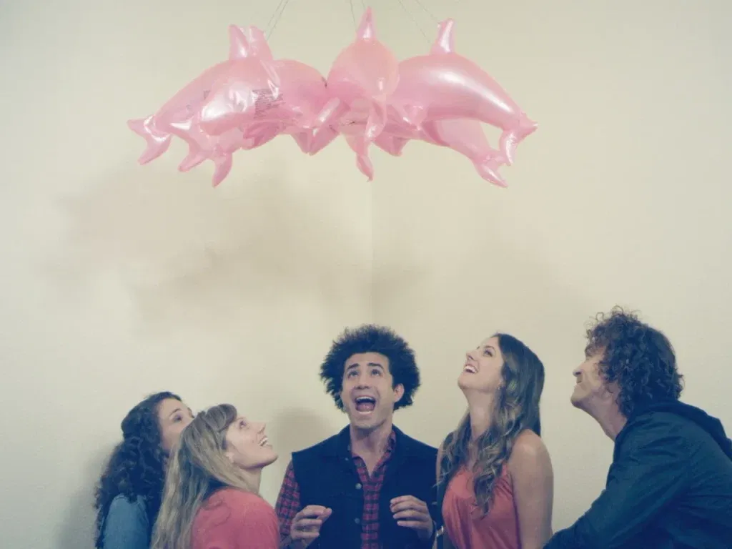 Characters from I Am Not a Hipster (2012) look up in wonder at a cluster of pink inflatable dolphin balloons hanging from the ceiling.
