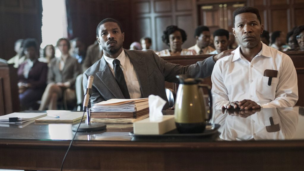 Michael B. Jordan as attorney Bryan Stevenson places a supportive hand on Jamie Foxx's shoulder as they stand together in a courtroom in Just Mercy.
