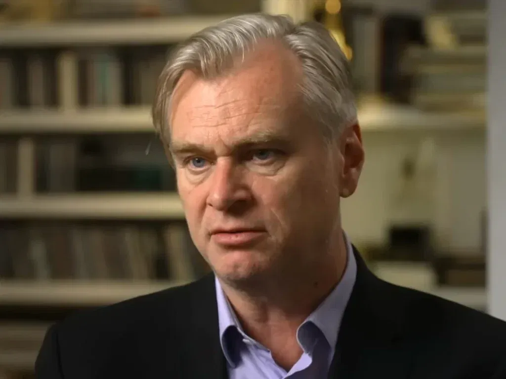 Christopher Nolan speaking in a close-up interview shot, wearing a dark blazer over a light blue shirt, with bookshelves blurred in the background.