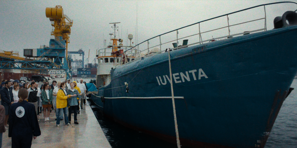 Volunteers and supporters gather beside the Iuventa rescue ship docked at a harbor before the vessel departs on its humanitarian mission in 23,000 Lives.