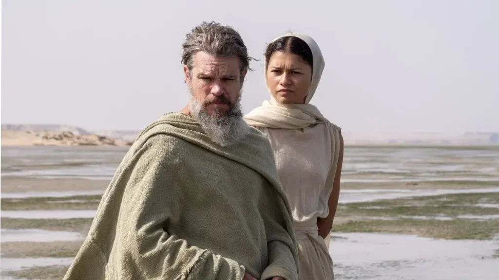 Matt Damon's Odysseus stands in the foreground while Zendaya's Athena watches from behind on a barren coastal landscape in Christopher Nolan's The Odyssey.