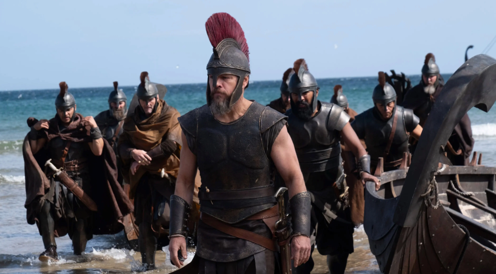 Matt Damon's Odysseus, clad in bronze armor with a red-crested helmet, leads a group of Greek warriors ashore from their ships in Christopher Nolan's The Odyssey.