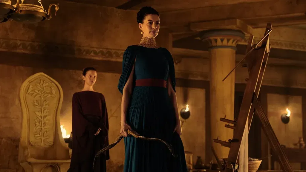 Anne Hathaway as Penelope stands holding a bow inside Ithaca's palace, with a young attendant behind her, in Christopher Nolan's The Odyssey.