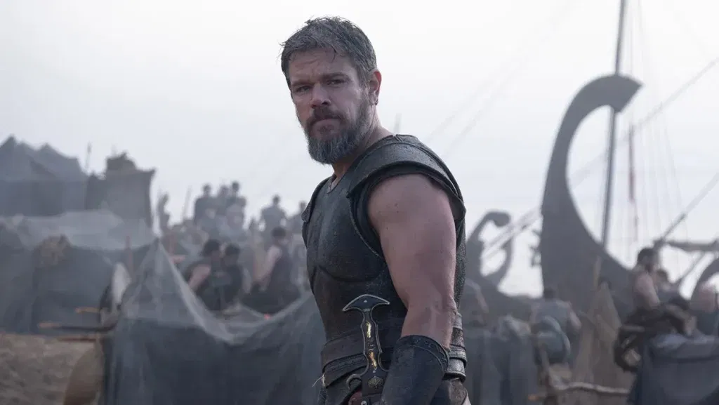 Matt Damon as Odysseus stands in a Greek military encampment in Christopher Nolan's The Odyssey, with soldiers, tents, and longships visible in the background.