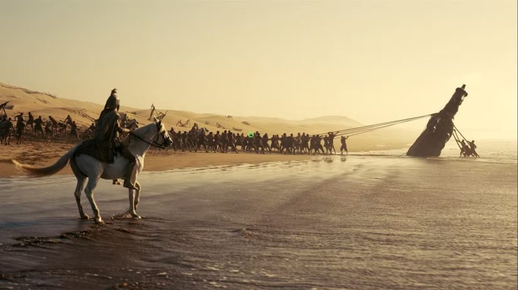 Greek soldiers haul a damaged wooden longship ashore at sunrise while a mounted warrior watches from the shoreline in Christopher Nolan's The Odyssey.
