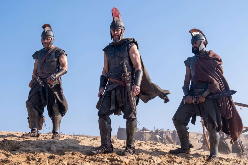 Matt Damon as Odysseus stands between two Greek warriors on a sandy battlefield in Christopher Nolan's The Odyssey, wearing bronze armour and a crested helmet.