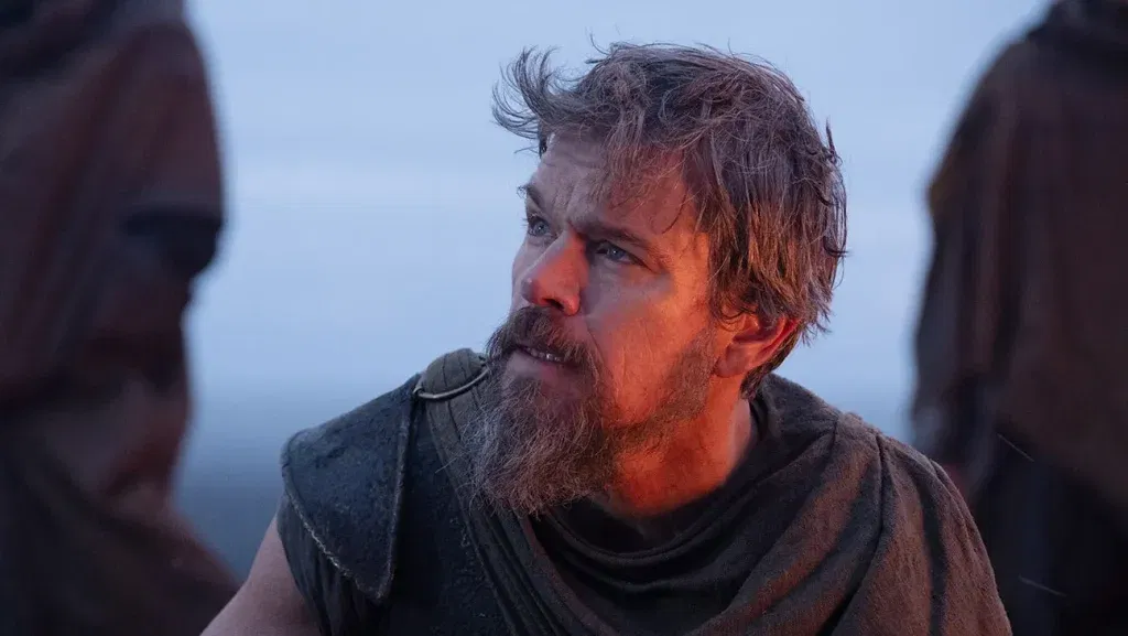 Close-up of a bearded warrior (Matt Damon) in weathered dark clothing looking upward with a determined expression as two hooded figures stand blurred in the foreground in The Odyssey.