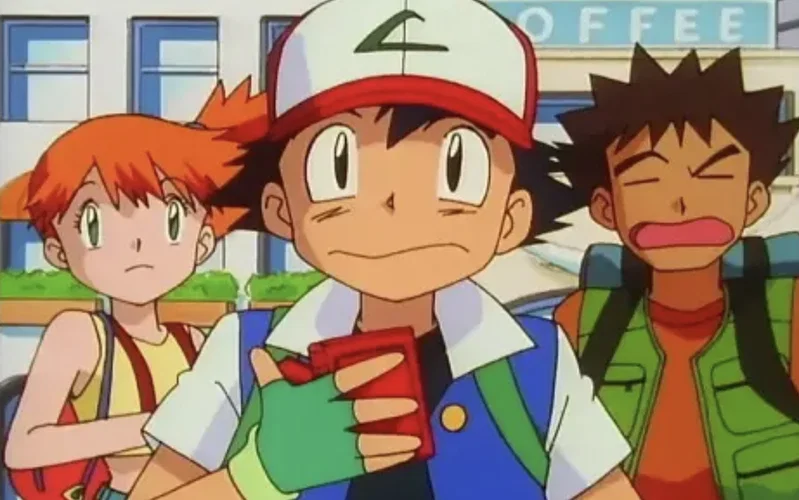 This scene features Ash Ketchum, Misty, and Brock from the Pokémon: Indigo League series. Ash is shown holding his original red Pokédex, a device used to catalog Pokémon data. The group appears to be standing in front of a coffee shop in the Kanto region.