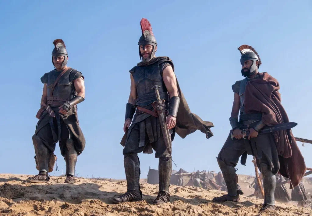 Three armored Greek warriors, led by Matt Damon in the center, stand on a sandy battlefield in Christopher Nolan's The Odyssey, with tents and soldiers visible in the background under a clear blue sky.