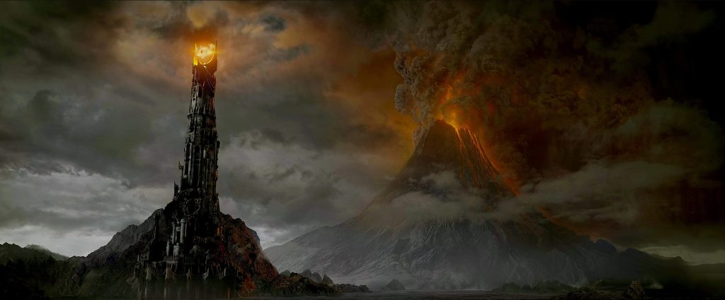 Barad-dûr rises beside the erupting Mount Doom, with the Eye of Sauron blazing atop the Dark Tower as smoke and ash fill the skies over Mordor in The Lord of the Rings.