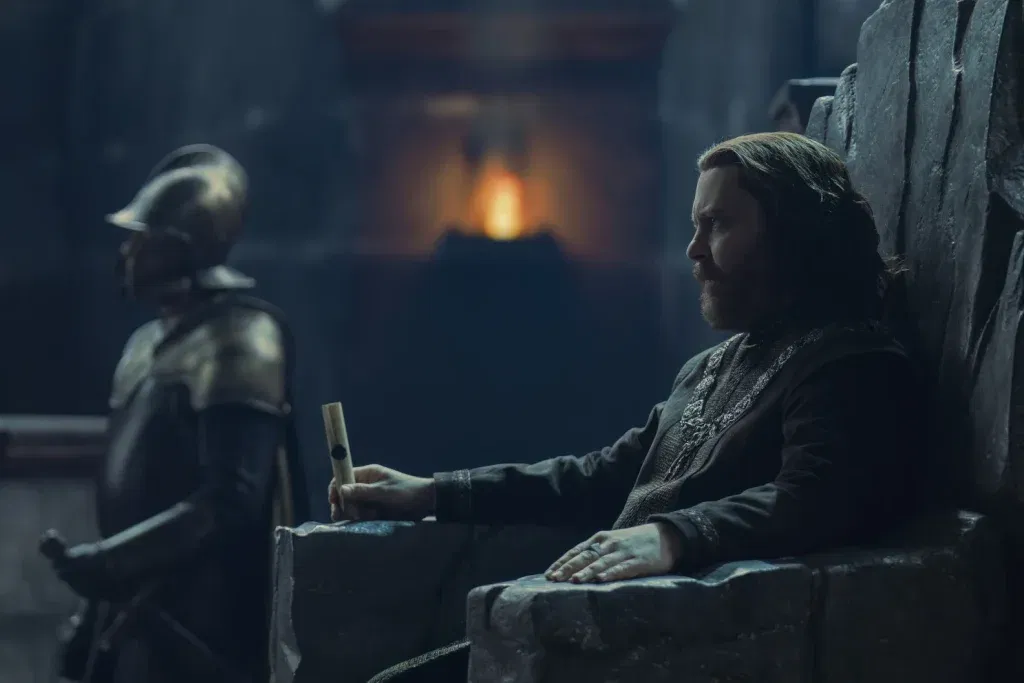 Lord Borros Baratheon sits on a stone throne holding a sealed scroll, an armored guard standing behind him in a firelit hall in House of the Dragon 