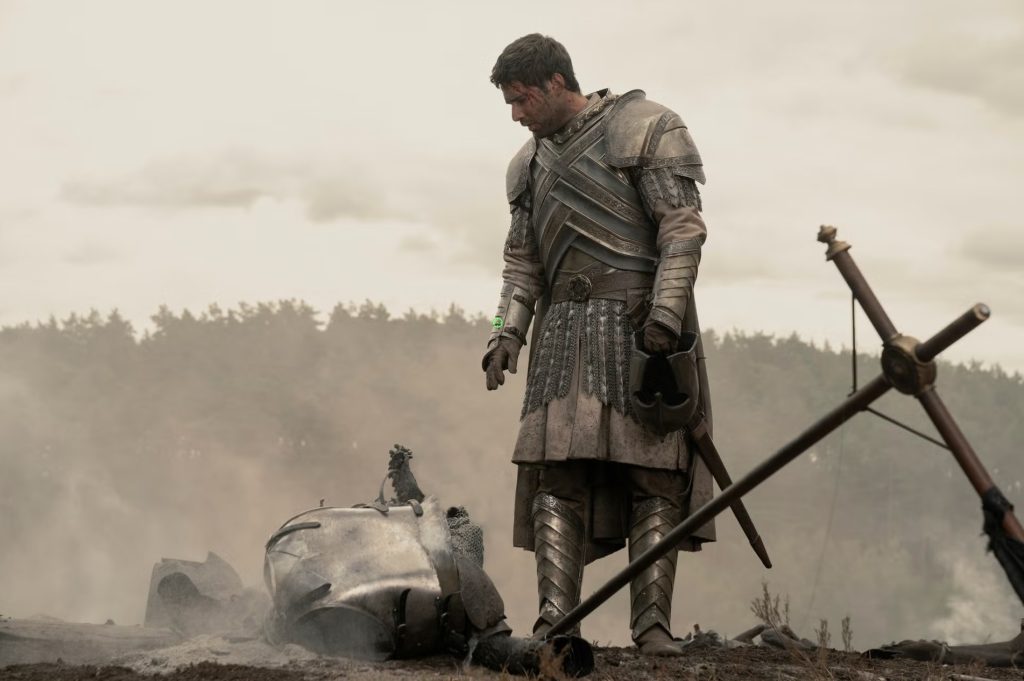Ser Criston Cole stands battered and blood-streaked in dusty full plate armor on a smoke-covered battlefield, helmet in hand beside broken armor, in House of the Dragon Season 3