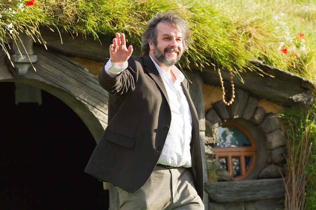 Peter Jackson waves while standing outside a hobbit hole at Hobbiton, wearing a dark blazer over a white shirt.