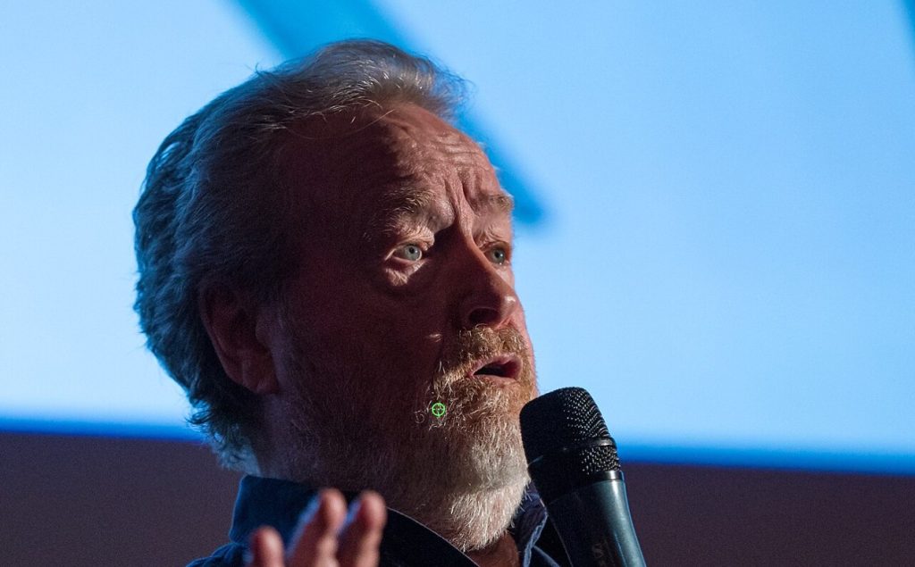 Ridley Scott speaks into a microphone while gesturing with his hand, photographed in close-up against a blue-lit backdrop at a public event.