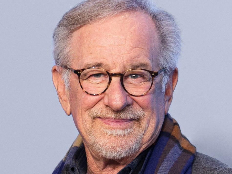 Steven Spielberg smiles at the camera, wearing round tortoiseshell glasses and a plaid scarf against a light blue backdrop
