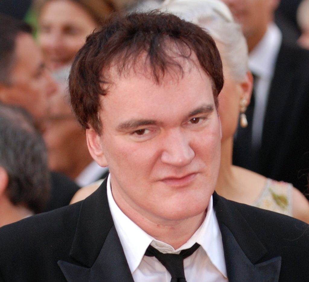 Quentin Tarantino in a black tuxedo and bow tie, photographed at a red carpet event with a crowd blurred in the background