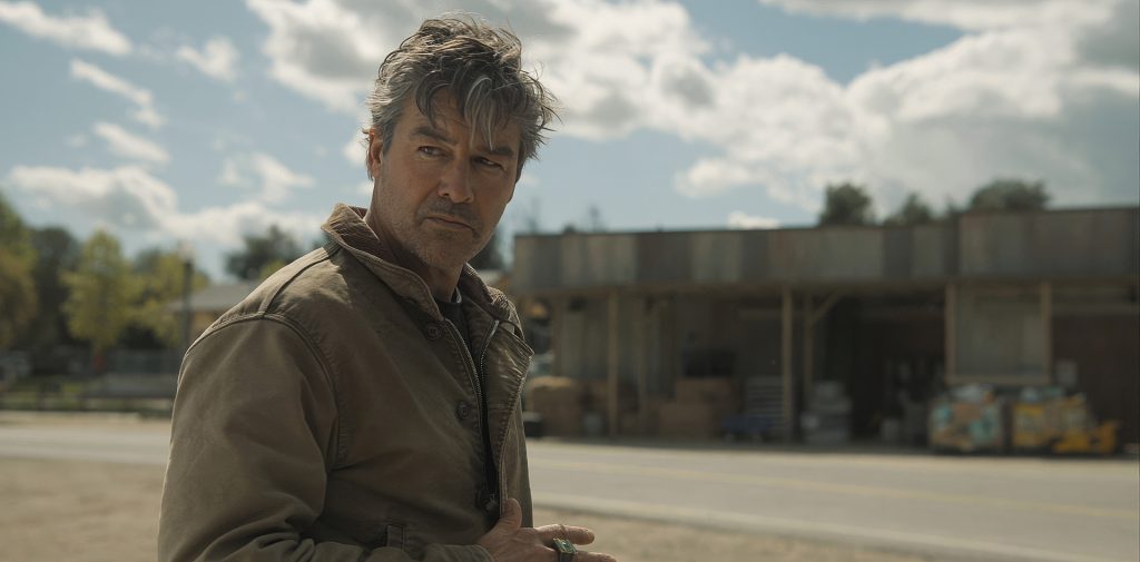 Kyle Chandler as Hal Jordan in HBO's Lanterns, standing on a small-town street in daylight wearing a weathered jacket, a green ring visible on his hand, looking off to the side with a wary expression.