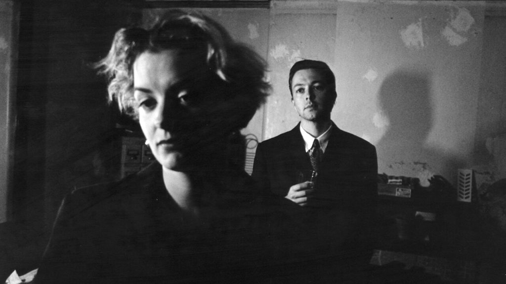 A woman stands in the foreground while a suited man watches from the background in a dimly lit black-and-white scene from Christopher Nolan's Following (1998).