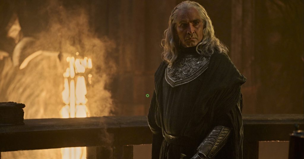  The Rings of Power Season 3, dressed in dark armored robes with long silver hair, standing before flames on a stone balcony.