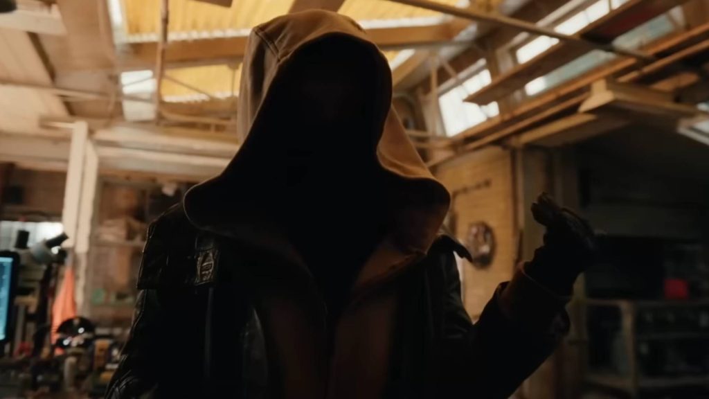 A hooded figure stands in a dimly lit industrial warehouse, their face completely hidden in shadow beneath a large hood. Wearing a dark leather jacket over a hooded sweatshirt and black gloves, the mysterious person gestures with one hand as warm light filters through the skylights above.