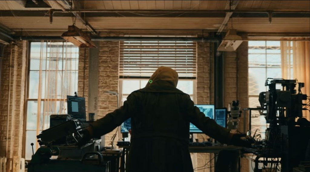 A hooded figure stands with their back to the camera inside a warehouse-like laboratory filled with computers, scientific equipment, and monitors. With both arms outstretched, the mysterious person faces a workstation bathed in daylight streaming through large industrial windows.
