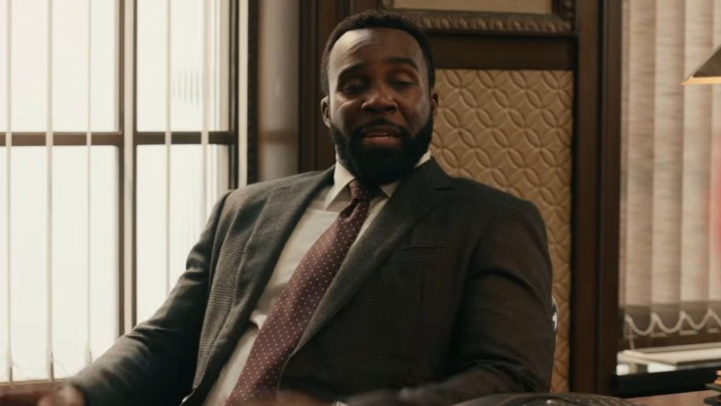 A man in a dark suit sits in an office chair during a conversation inside a well-appointed office. Sunlight streams through tall windows behind him as he speaks with a composed expression.