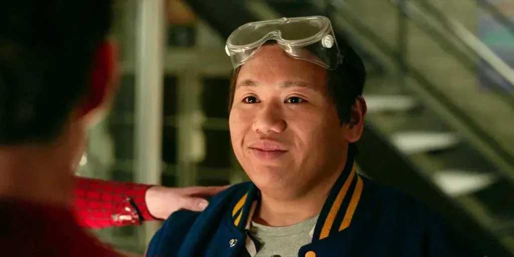 Ned Leeds smiles warmly while wearing safety goggles pushed up on his head and a blue varsity jacket. Spider-Man, seen from behind in the foreground, rests a hand on Ned's shoulder during an emotional reunion.