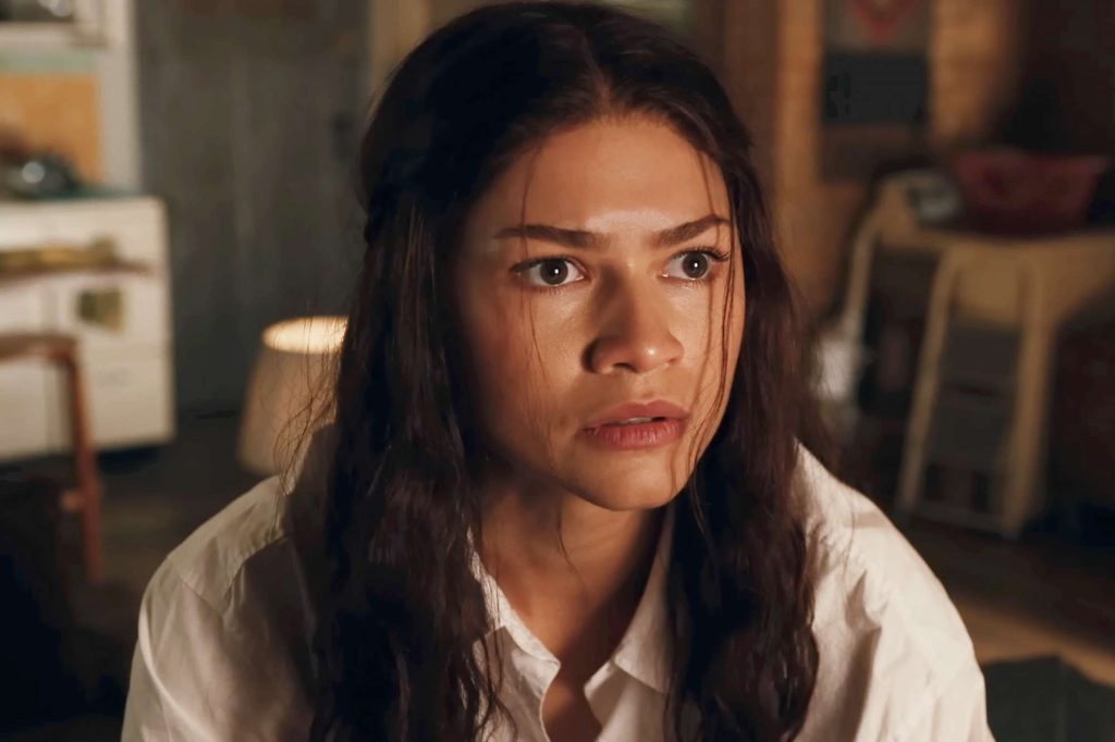 MJ looks ahead with a worried expression inside a warmly lit apartment. Wearing a white button-up shirt, she appears deep in thought as the softly blurred room behind her creates an intimate atmosphere.