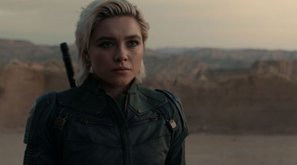 Yelena Belova stands in a rocky desert landscape wearing her black tactical suit. With a serious expression and a weapon strapped across her back, she looks toward something off-camera under an overcast sky.