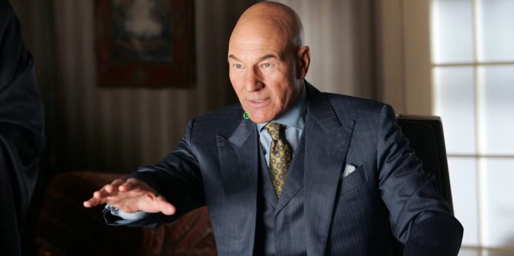 Professor Charles Xavier sits at a desk wearing a blue pinstripe suit and patterned tie. He gestures with one hand while speaking in a warmly lit office, his expression calm and attentive.