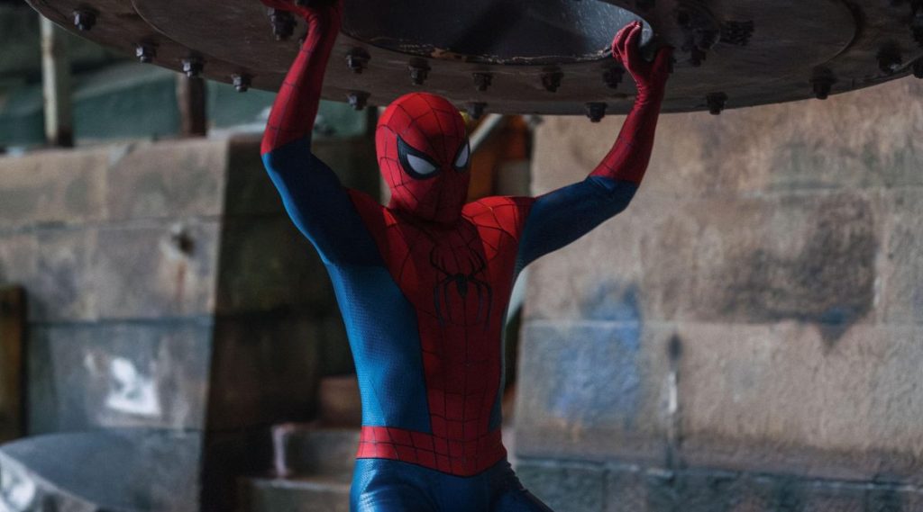 Spider-Man braces himself beneath a massive circular metal structure, holding it overhead with both hands inside an industrial facility. Wearing his red-and-blue suit, he strains to keep the heavy object from collapsing.