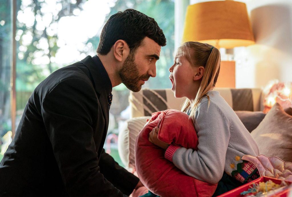 Roy Kent leans in to speak with his niece Phoebe during a heartfelt moment in Ted Lasso, as she clutches a pink cushion inside a warmly lit living room.
