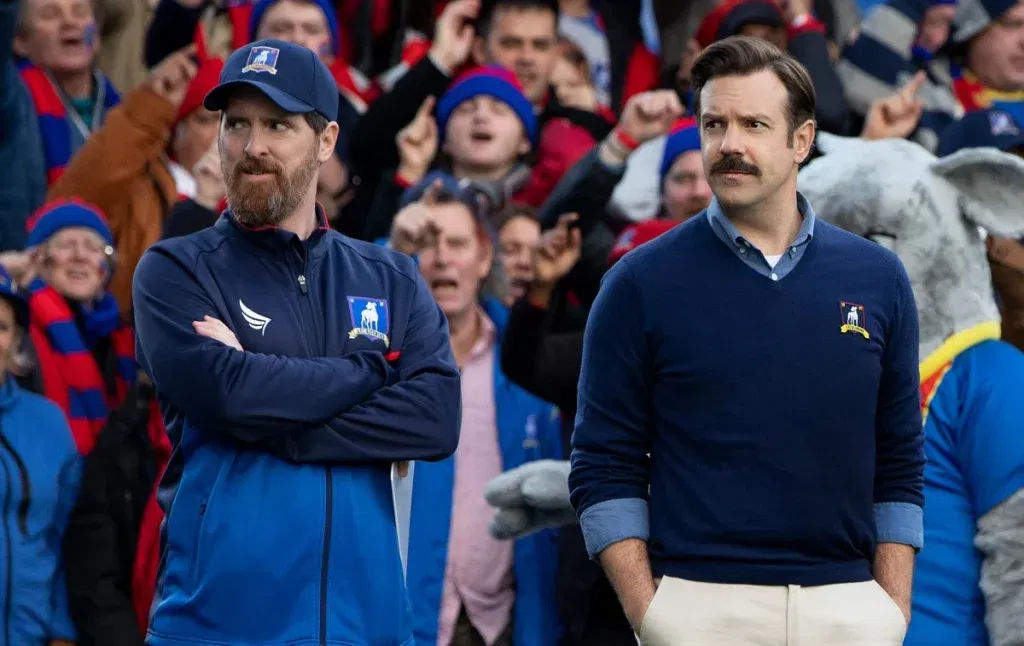 Ted Lasso and Coach Beard stand on the sidelines during an AFC Richmond match, watching the action unfold with contrasting expressions in Ted Lasso.